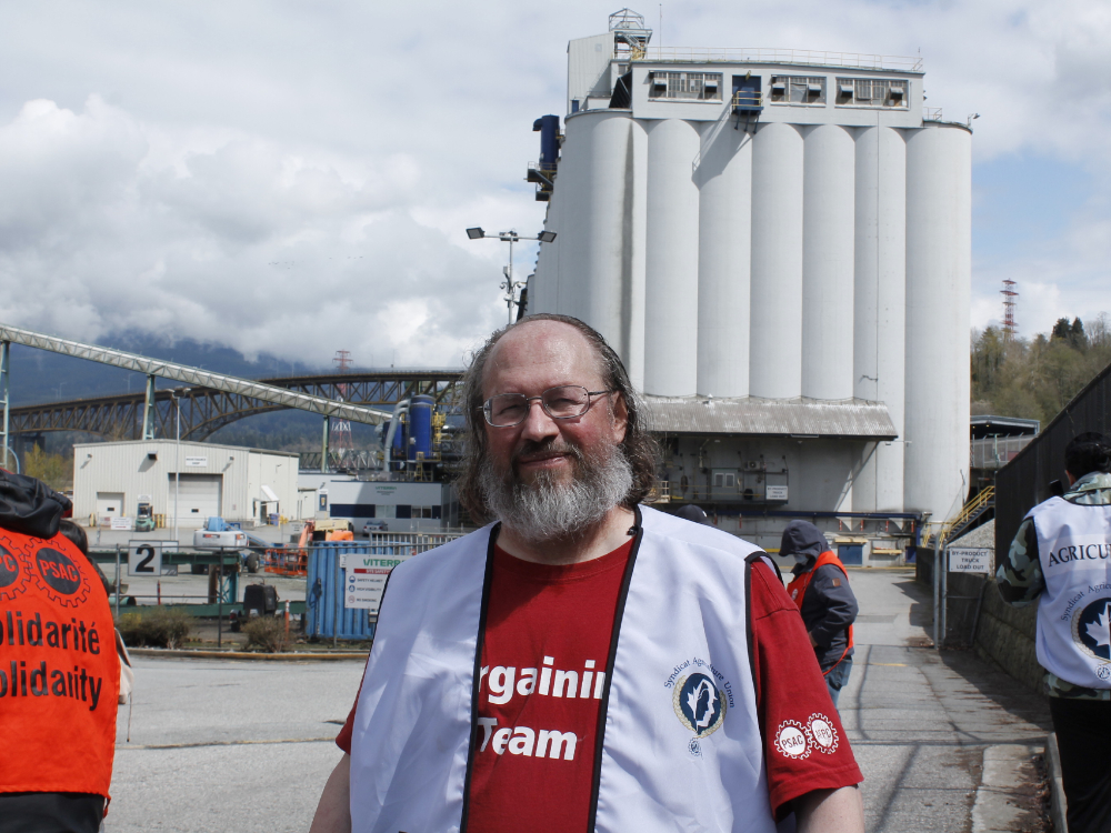 A bearded man wearing a red T-shirt and grey vest stands in front of a concrete grain elevator.