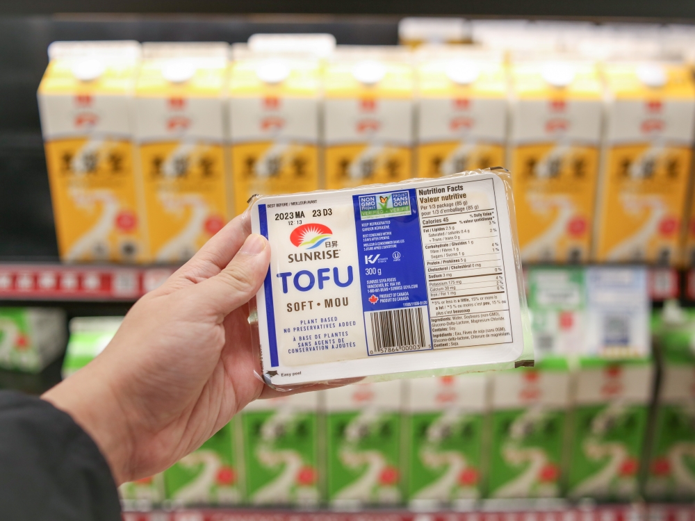 A hand holds a package of Sunrise soft tofu in front of a fridge of soy milk.