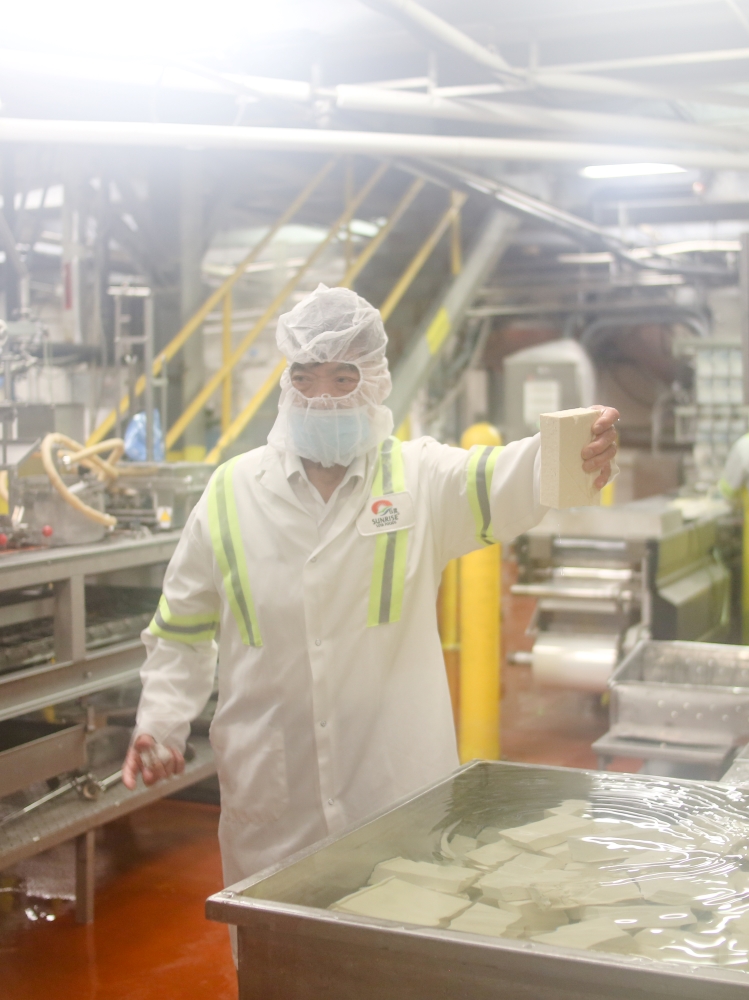 A Chinese man in his 80s holds a block of tofu that he just plucked from a cooling bath. He is in a production facility surrounded by machines. The camera lens is foggy from the steam.