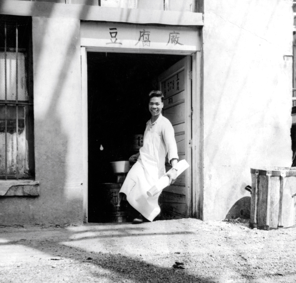 An old black and white photo of a building on a sunny day. The words “tofu factory” are written in Chinese characters above a doorway. A smiling man is standing there in an apron.