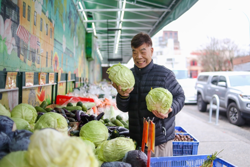 An 88-year-old with a big smile rearranges cabbages at the street-front of a green grocer.
