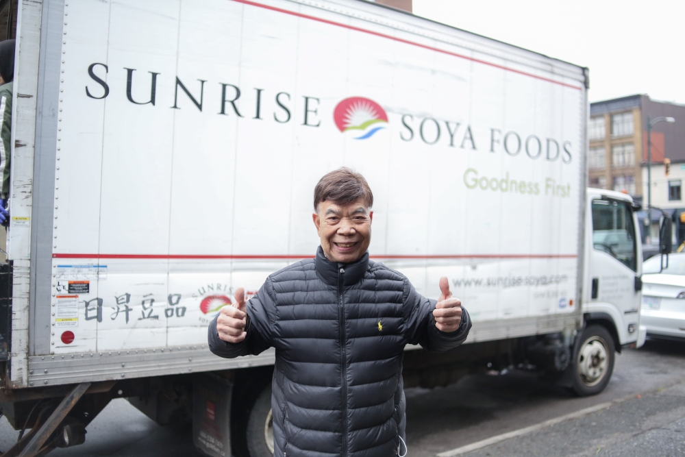 An 88-year-old with a big smile holds two thumbs up in front of a truck with the letters “Sunrise Soya Foods.”