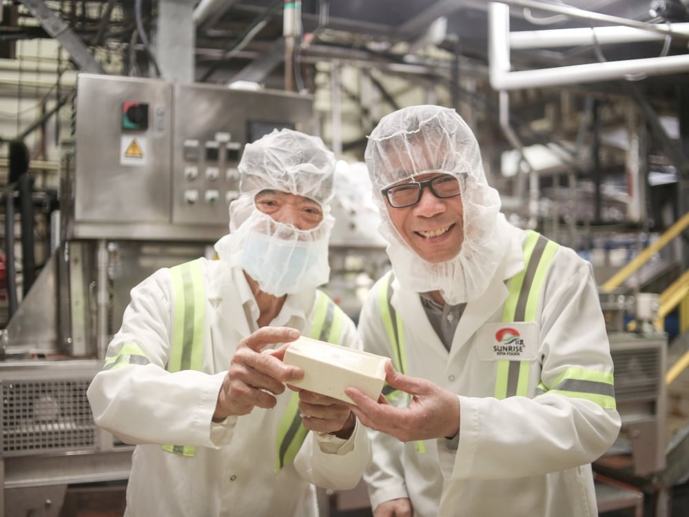 Two Chinese men in white suits and food safety hair nets smile at the camera while standing in an industrial food processing facility. Both their hands hold a single block of white tofu.