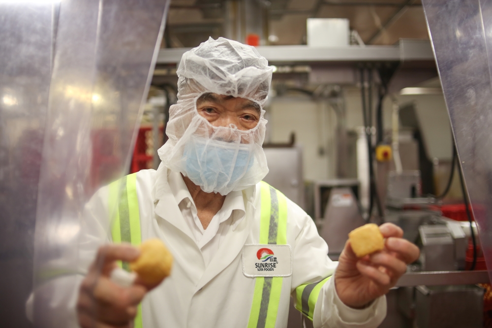 A Chinese man in his 80s wearing a mask and a hooded net walks through a curtain holding two tofu puffs.