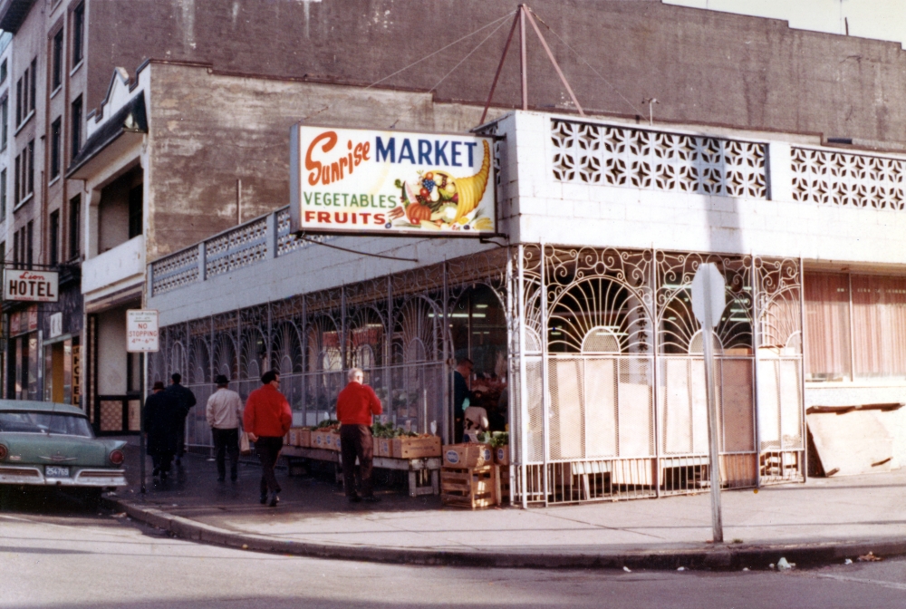 An old photo of a small market on the corner of a city block. The sign, which reads Sunrise Tofu, has the log of a cornucopia of fruits and vegetables.