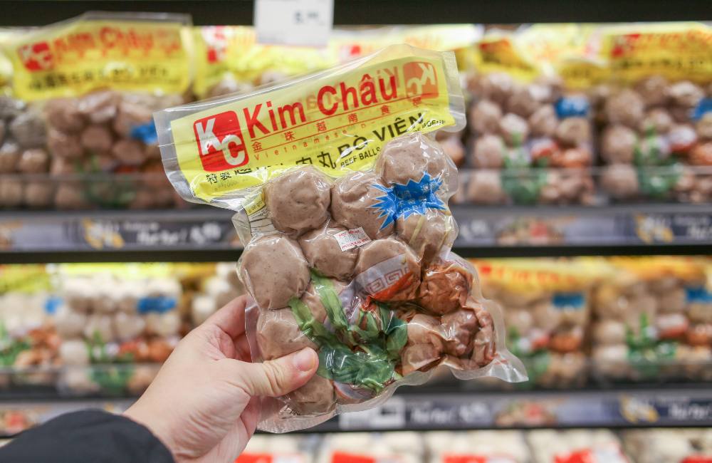 A hand holds a pack of vacuum-sealed beef balls in front of a supermarket fridge. At the top in red letters is the brand “Kim Chau.”