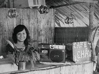 A photo from the late-1970s showing a woman smiling behind the counter of a tropical-looking stall. There is a tape player on the counter beside a sign that reads Cafe Chau.