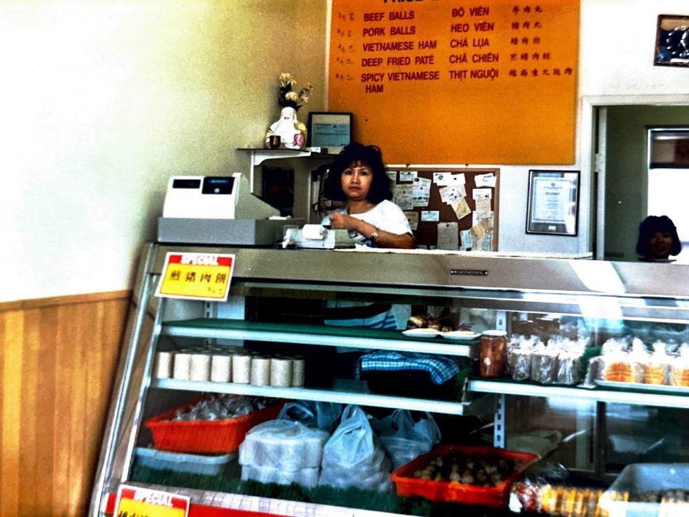 A photo from the late-1980s showing a woman at a deli counter with packages of Vietnamese ham and a menu behind her. 