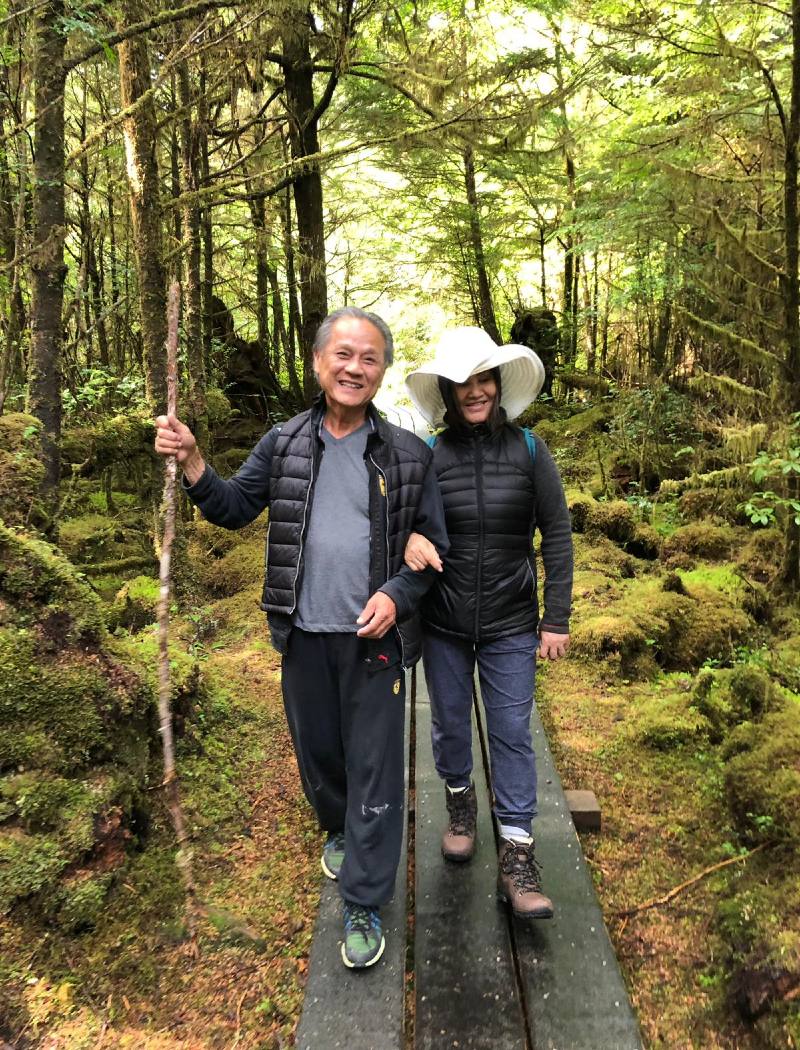 An adult daughter walks with her elderly father on a forest trail. She’s holding onto his arm and they’re both smiling.