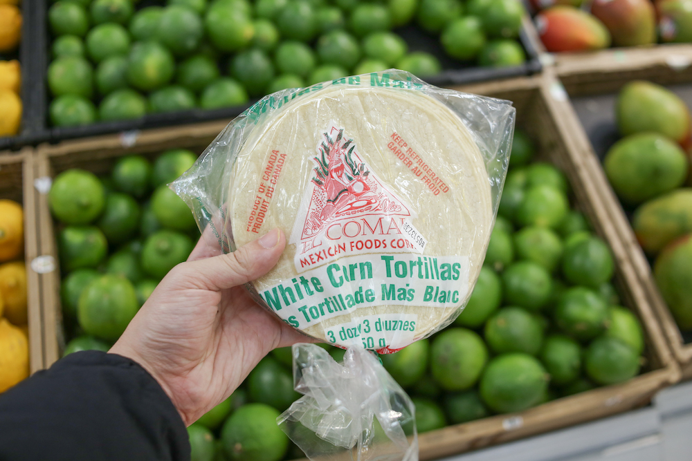 A hand holds a pack of El Comal brand tortillas in front of a freezer.