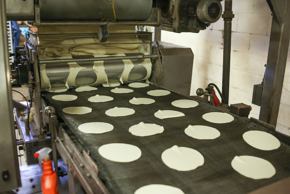 A production line with four lines of tortillas coming off the conveyer belt. You can see the dough from which they are cut.