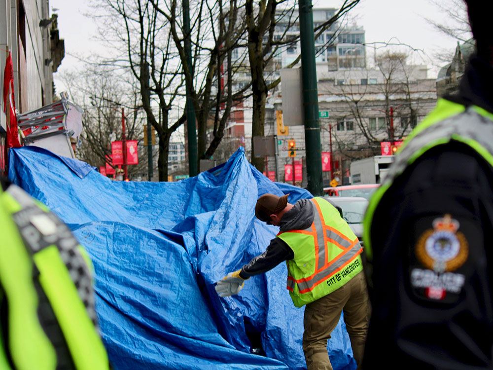 A city worker in a neon yellow utility vest and backwards ball cap has their back turned to the camera. They are reaching towards a large tent structure covered by a bright blue tarp in an urban setting. In the blurry foreground, the shoulders of Vancouver police officers are visible, their police insignia visible on the shoulder of one person to the right of the frame.