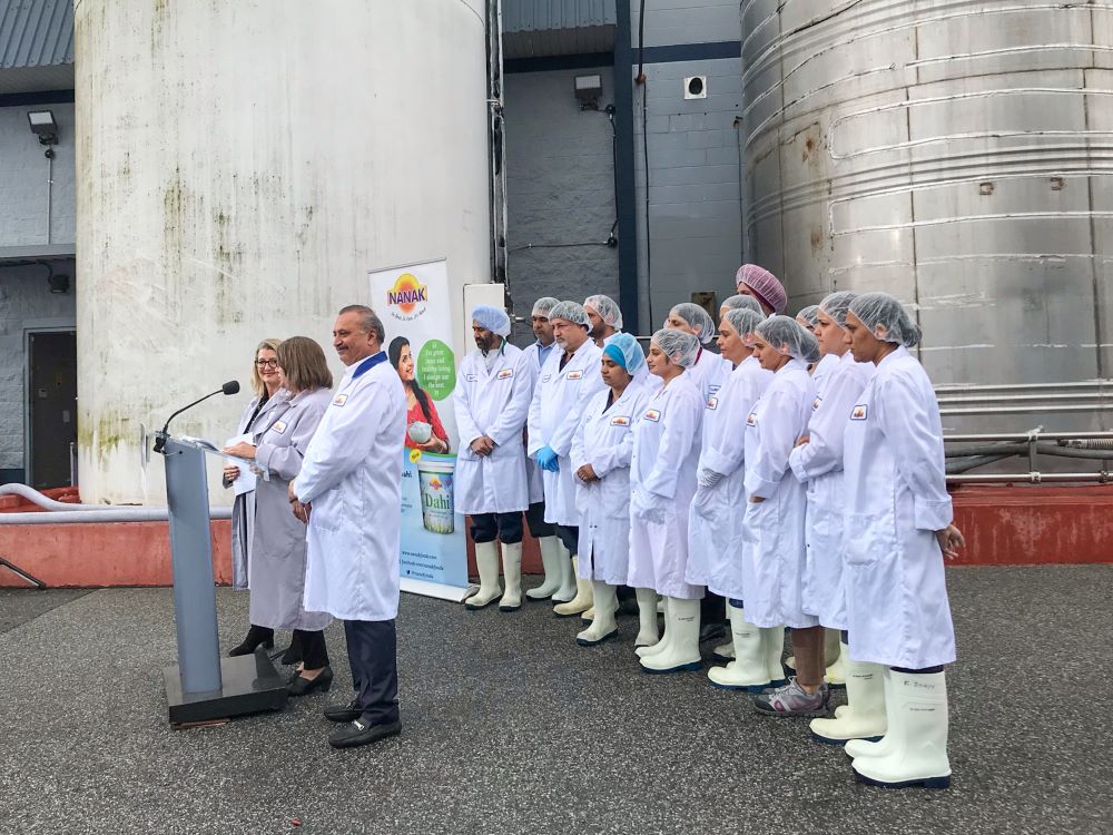 Three dignitaries stand at a podium while a crowd of factory staff in white attire stand behind. There is a factory in the background with large stocks.