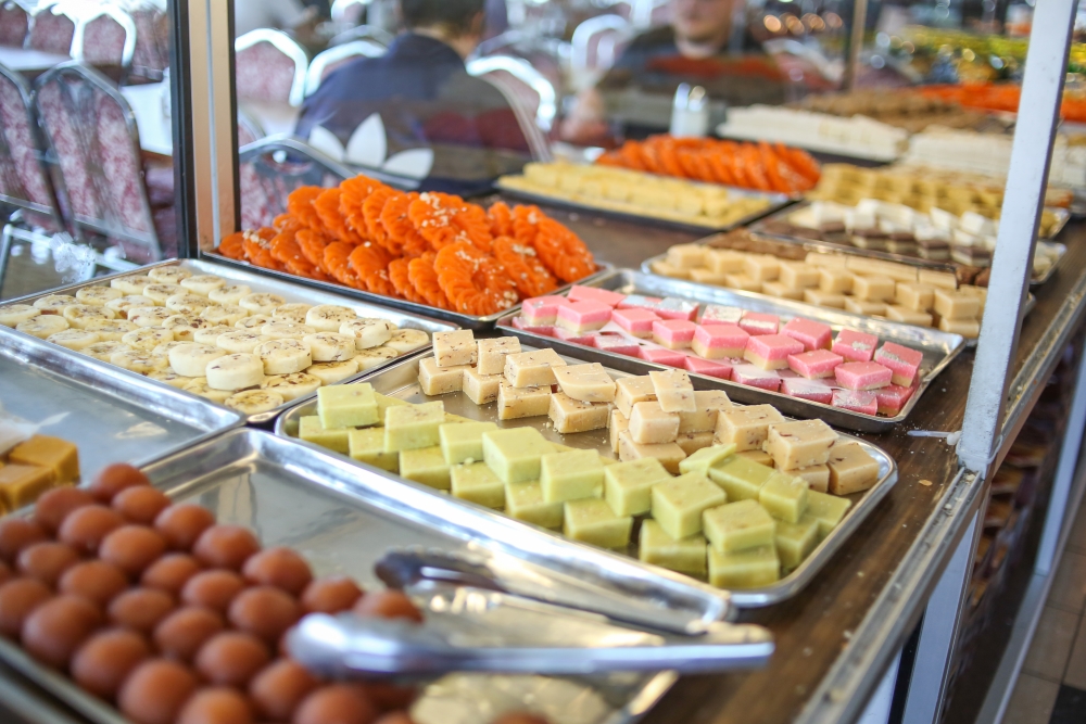 Orange, pink, cream, light green Indian sweets in a buffet display case, with tongs at the ready.