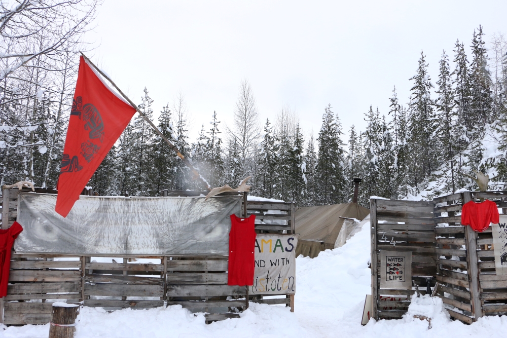 A high wooden fence hung with red dresses and a red flag encloses a camp, which is obscured by the fence.