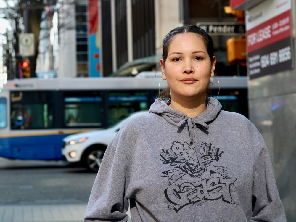A person with hoop earrings and a grey hoodie looks at the camera, as traffic passes behind.