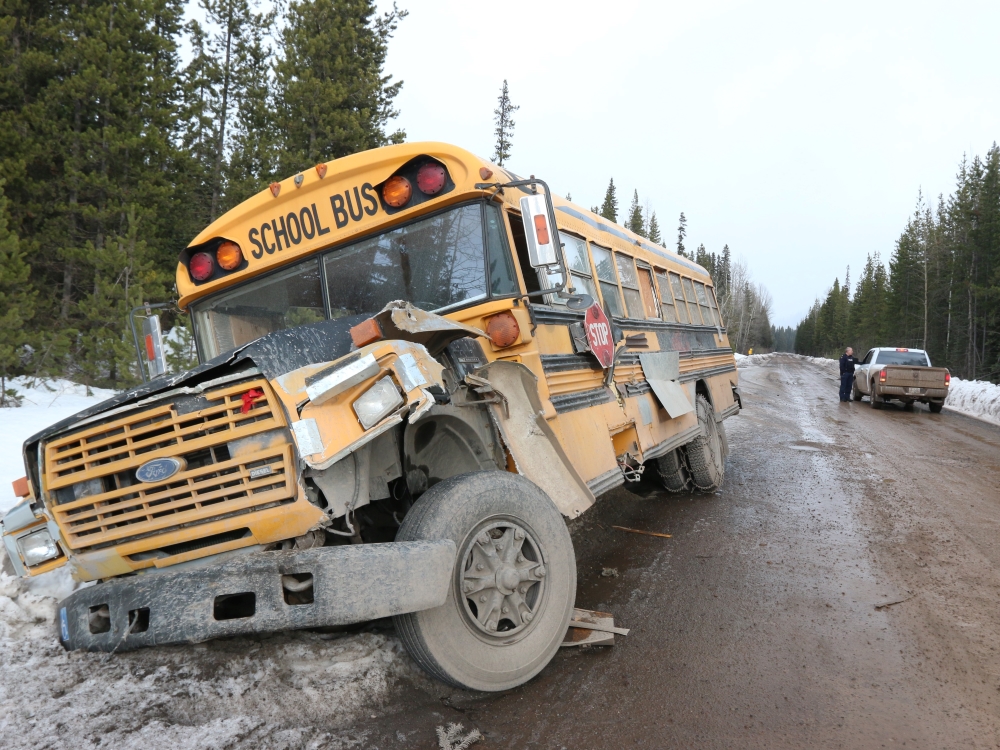 An old yellow school bus is at the left of the photo, pulled into a snowy ditch on a muddy road. Just beyond, at right of the photo, a man in uniform speaks with the driver of a pickup truck.