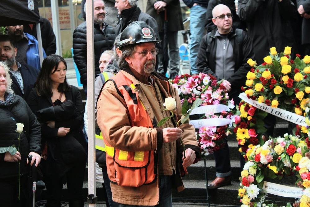 A person in a black hard hat and safety vest holds a single white rose. Flower wreaths are to his left. People are gathered behind.