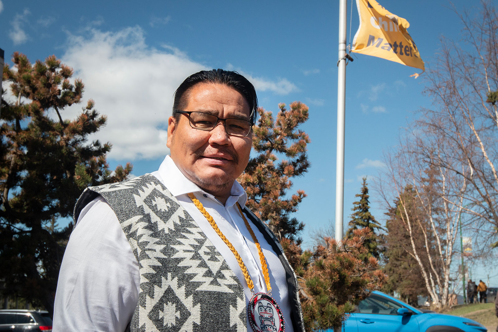 A man with dark hair and glasses stands below an orange flag and blue sky. He is wearing a vest with traditional Indigenous designs and a beaded pendant.