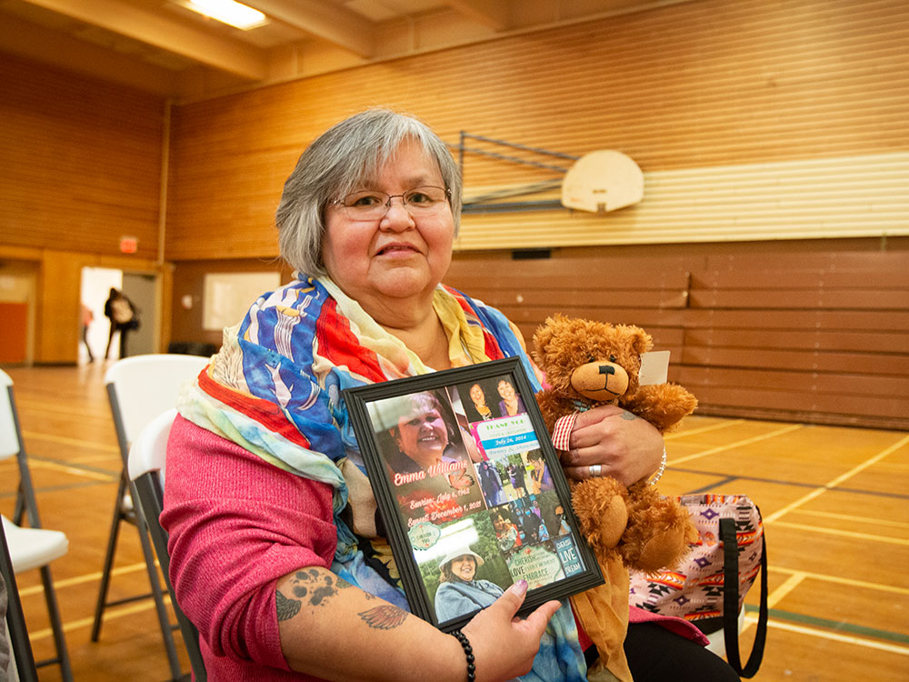 A woman with short, greying hair sits inside a wood-panelled gymnasium holding a teddy bear in one hand and photo frame in the other. She is wearing a pink shirt and brightly coloured scarf. Photo by Amanda Follett Hosgood.