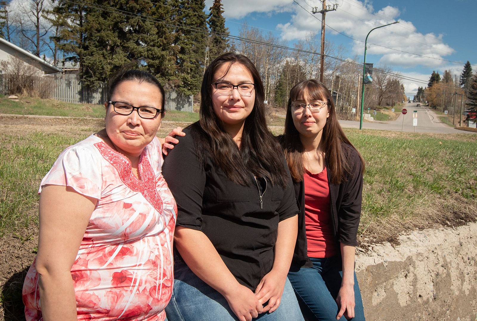 Three women, all with dark hair and wearing glasses, lean against a low wall with their arms around each other under a blue sky.