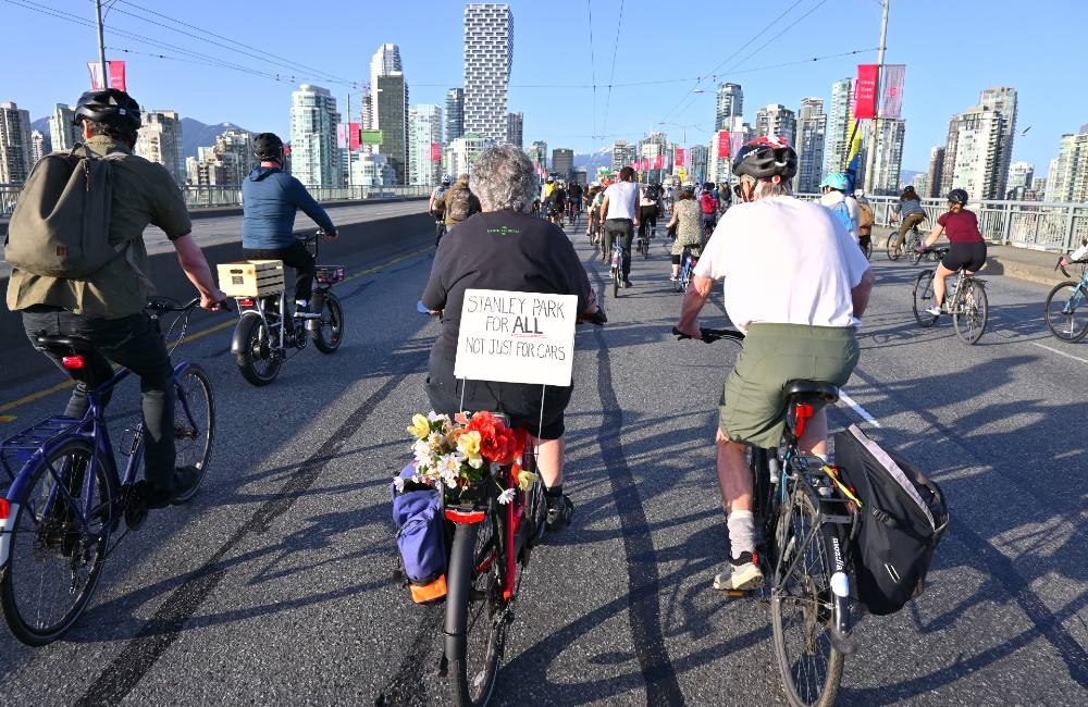 An older woman in a black t-shirt rides her bike towards downtown Vancouver with a large procession of other cyclists. She has flowers and a pannier attached to the rack of her bike. A sign also attached reads “Stanley Park for all, not just for cars.” 