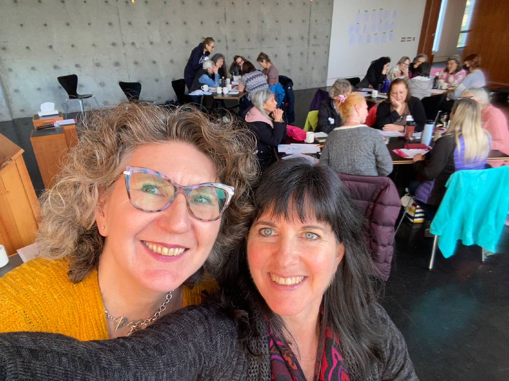 Two women take a selfie in an indoor space where people are gathered around tables, talking. On the left, Christa Ovenell has curly hair and glasses; on the right, Reena Lazar has dark hair and light blue eyes. 