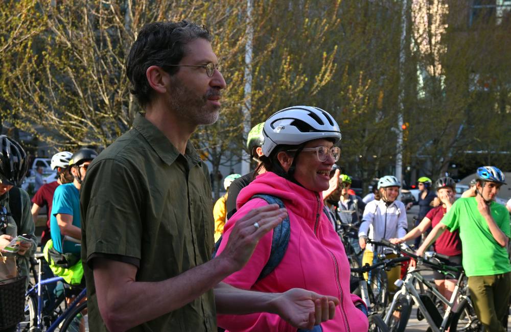 A man with a short beard and wearing a green shirt and glasses smiles and claps as he looks on. Beside him a person with pink glasses and in a bright pink jacket and a white helmet laughs. Other cyclists stand around also smiling and laughing.