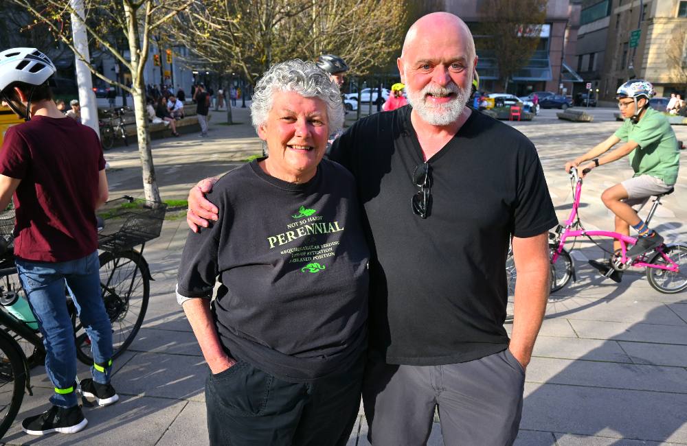 Two older people embrace at their sides surrounded by cyclists. The woman standing to the left has short grey hair and wears a black t-shirt that reads ‘Not so hardy: Perennial.’ The man to the right is bald, has a grey beard and has bright blue eyes.