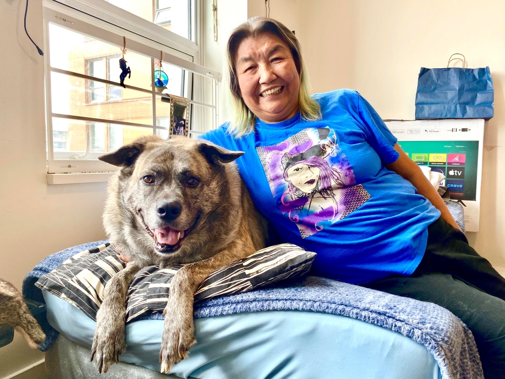 Marilyn Johnson is an Indigenous woman wearing a royal blue T-shirt with a purple and black illustrated graphic of her granddaughter. She is smiling at the camera, and she is leaning towards her large dog, Papoose, to the right of the frame; they are both reclining on a bed with a light blue sheet.