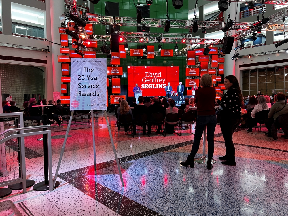 People gather in an atrium. A sign reads ‘The 25 Year Service Awards.’ Screens surround a stage. On the main screen, it reads, ‘David Geoffrey Seglins.’ 