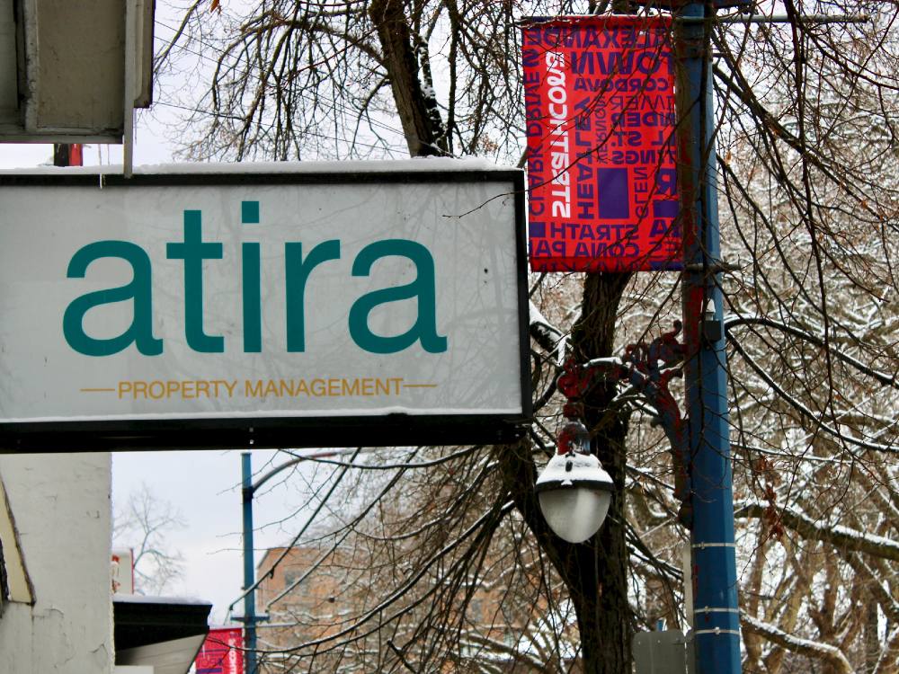A white sign reading 'Atira Property Management' juts out onto a street in winter. Trees and a lamppost covered in a slight coating of snow surround it. A red flag on the lamppost reads 'Strathcona,' with the names of streets in the Vancouver neighbourhood around it.