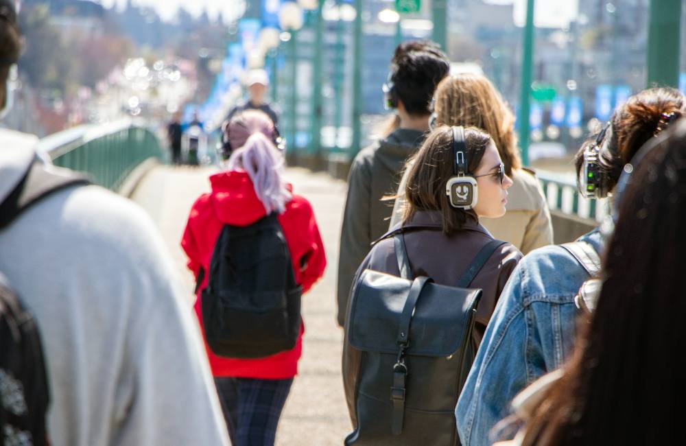 A group of young adults walks across the Cambie Bridge, memorable for its wide sidewalk and green railings, in Vancouver on a sunny day. Members of the group are wearing headphones and are walking with their backs turned to the camera.  