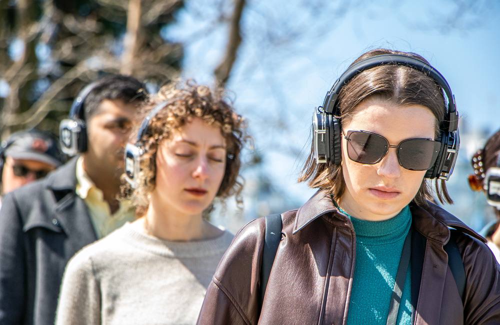 Four people participate in a ‘Walk with Me’ walk in Vancouver on a sunny day. They are wearing headphones and one person with curly hair and a grey sweater is closing their eyes. Another person in front of them is wearing sunglasses and a brown leather jacket.