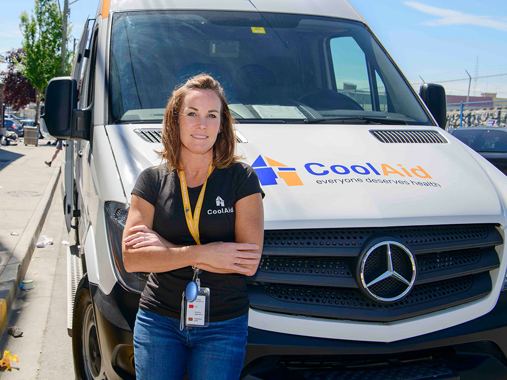 A woman with shoulder-length brown hair is wearing a black T-shirt and standing with her arms crossed in front of a white Mercedes Sprinter van on a sunny day. The blue and white logo for the Victoria Cool Aid Society is on the hood of the van.