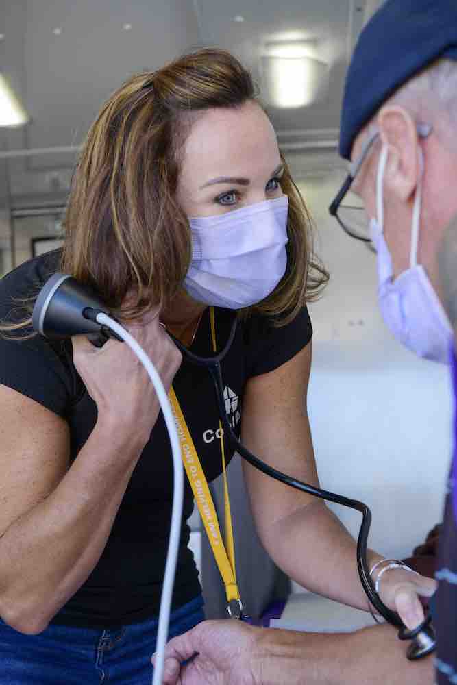 Karen Lundgren uses a stethoscope to with a patient in a mobile health clinic. She is wearing a black T-shirt and a purple medical mask. The patient’s face is not visible in the frame; they are sitting to the right of the frame and wearing a toque and purple medical mask.