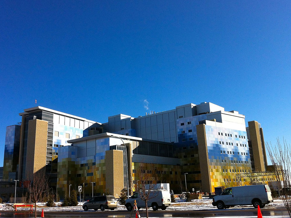 A massive hospital complex sits under crystal blue sky. Snow covers the ground, and trucks cars and pylons dot the pavement.