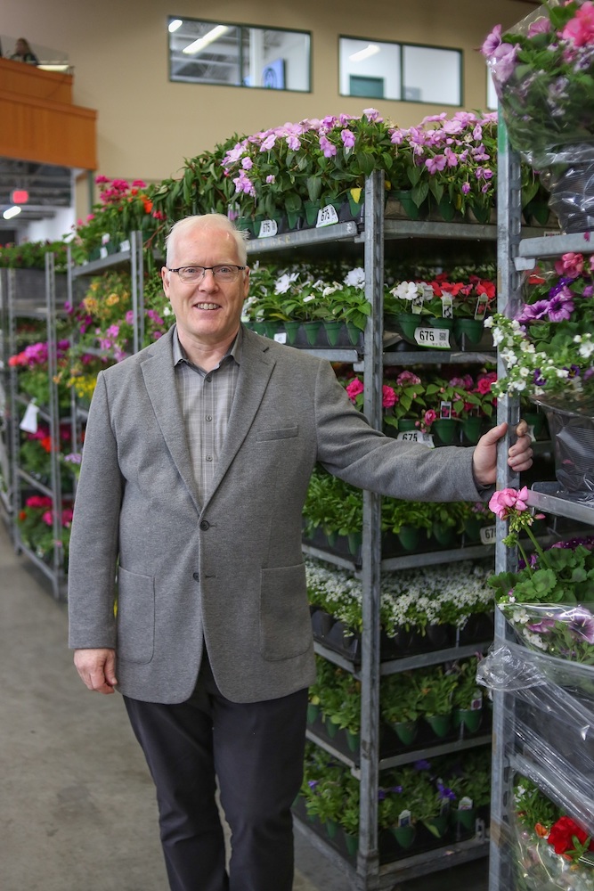 A man in a suit stands in a warehouse amidst racks of flowers.