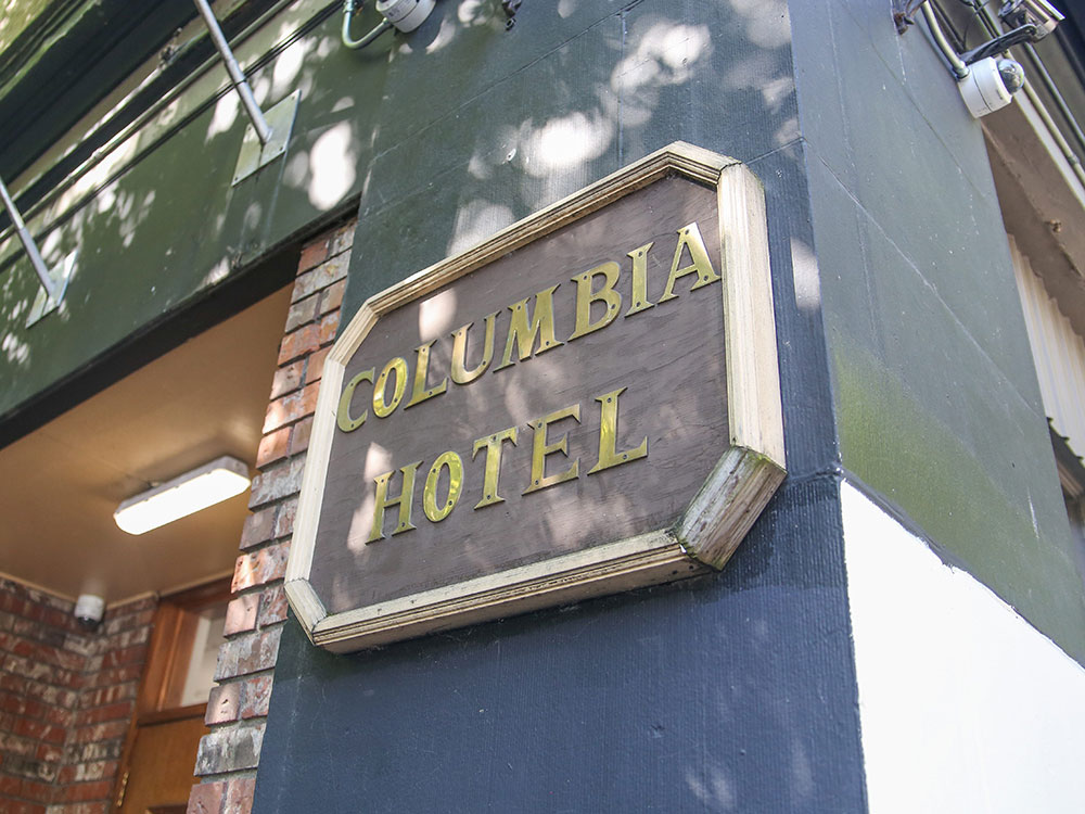 The exterior of 303 Columbia St. in Vancouver’s Downtown Eastside features a wooden plaque bearing gold capital letters that read “Columbia Hotel” against a painted blue, green and white exterior. A brick alcove and doorway is visible to the left of the frame. It’s a sunny day.
