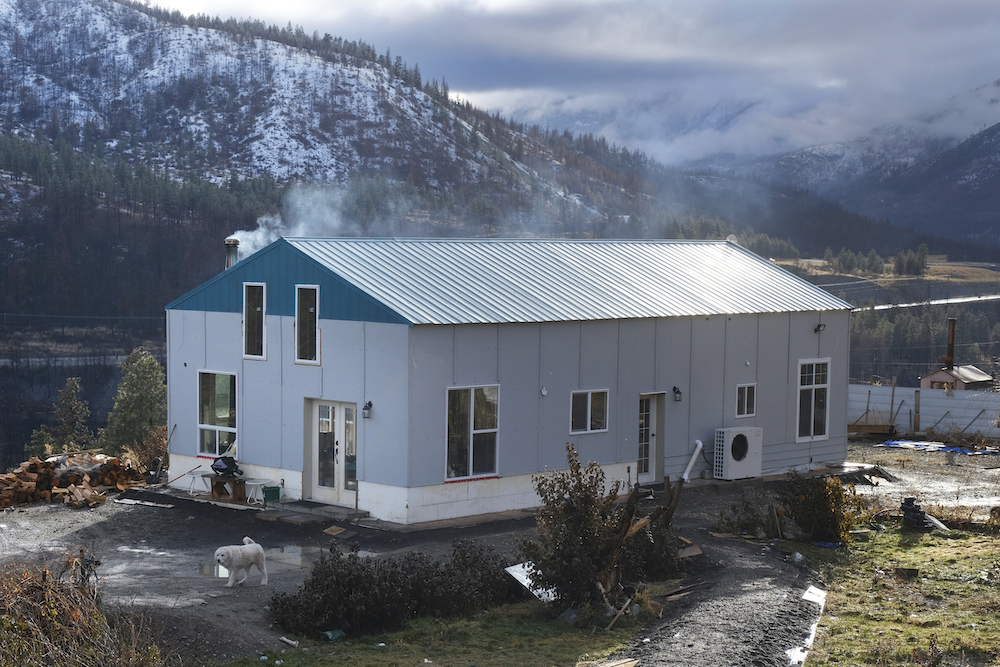 A blue and white house sits in a valley. Snow is on the surrounding mountains and steam is blowing off the home's roof. 