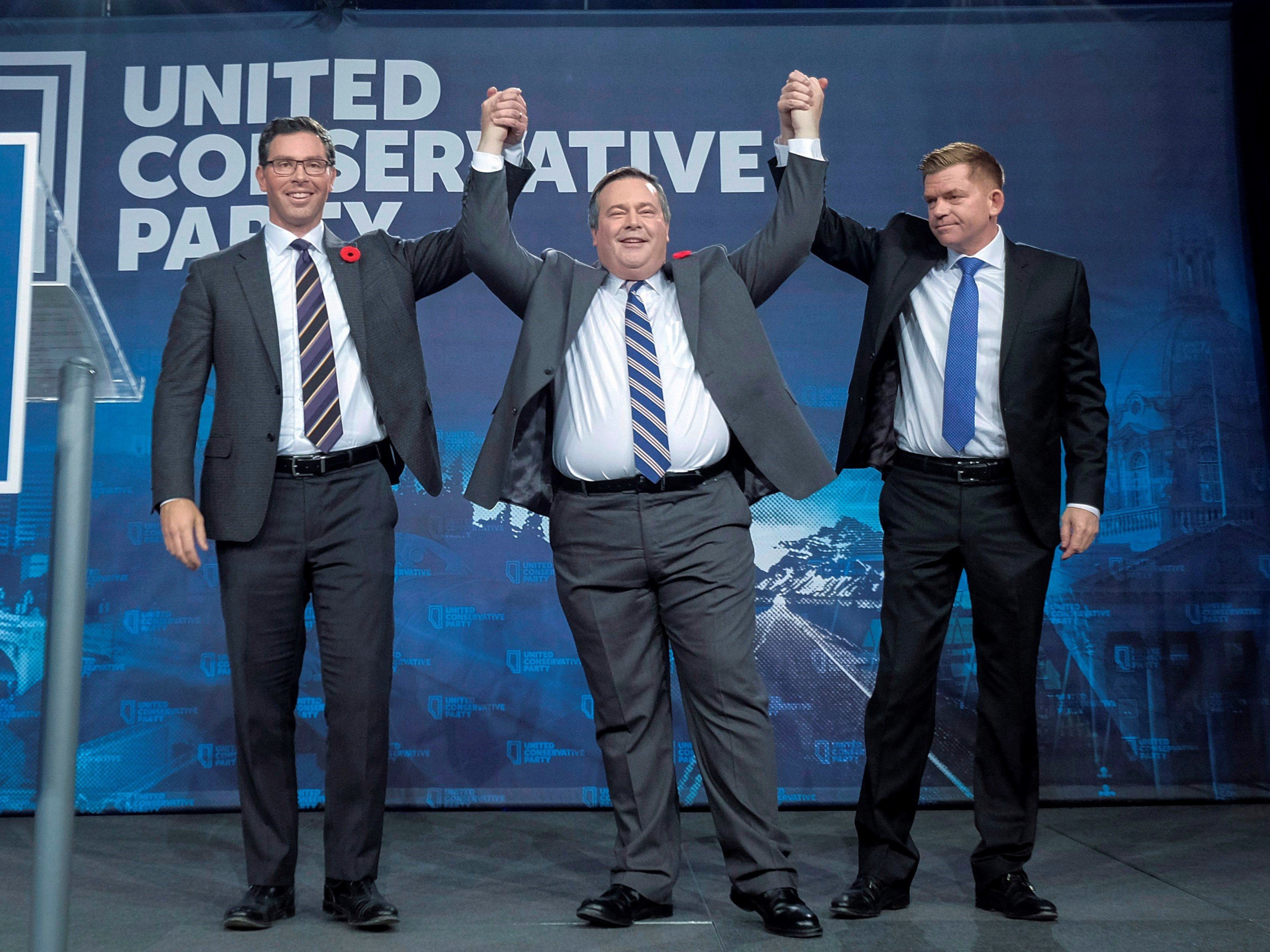 Three men stand on a stage wearing suits and ties. They are holding hands, with their hands in the air triumphantly. 