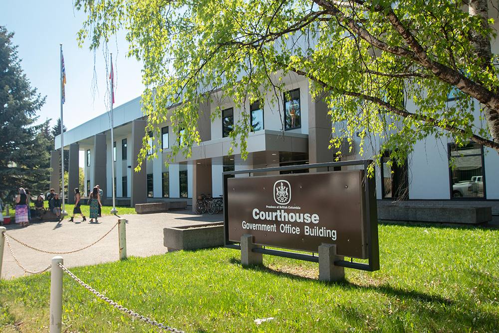 A brown sign that says “courthouse government office building” stands in front of a white and beige institutional looking building. There is blue sky and a birch tree overhanging the sign.