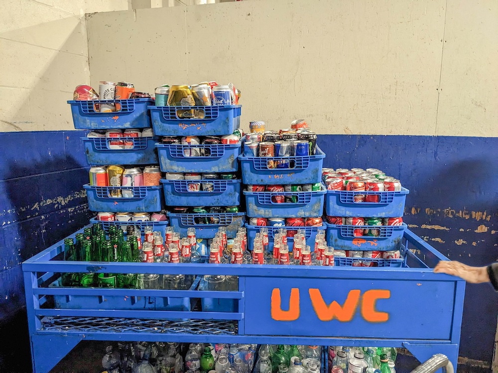 At a recycling depot, a trolley full of trays of recyclables, organized by cans and bottles. A hand belonging to a senior is pulling the trolley.