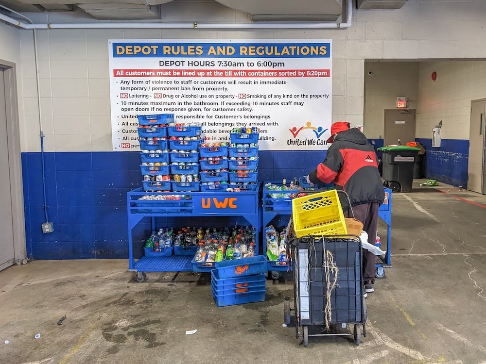 A man wearing a puffy red jacket organizes a trolley full of trays of cans and bottles. Behind him is a wall with a sign that has violence won’t be tolerated at the depot.