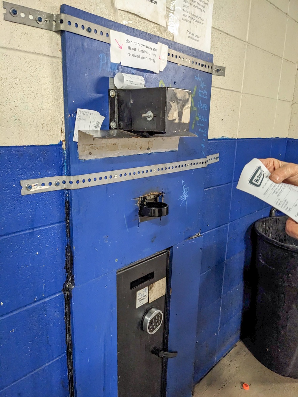 A man holds a receipt on the right side of the image. On the left is a wall with a machine embedded in it. At the base of the machine is a safe.