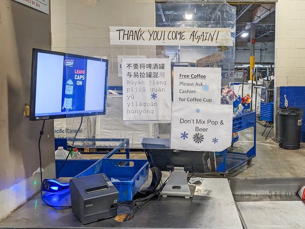 A cashier at a recycling depot with a hanging plastic screen. On it are signs in English and simplified Chinese that say not to mix pop cans and beer cans. Another sign says customers are welcome to free coffee.