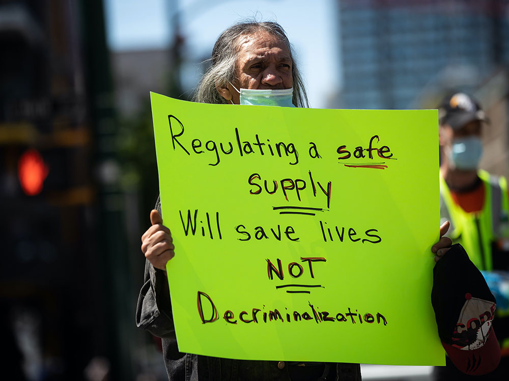 A person wearing a COVID mask holding a green sign that says “Regulating a safe supply will save lives. Not decriminalization.”