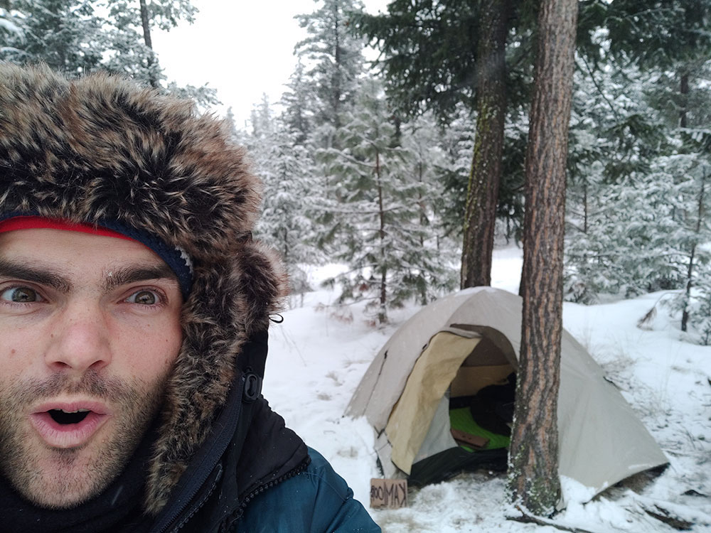 Timothy, wearing a hat and a lot of winter layers, takes a selfie at a snowy campsite.