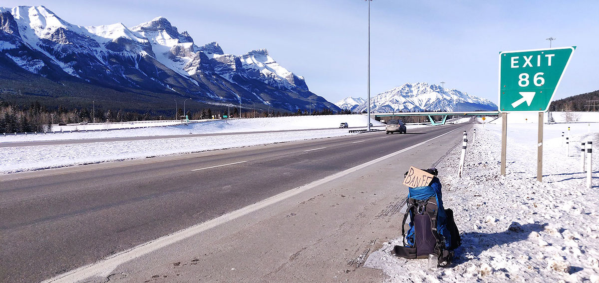 A cardboard sign with the word BANFF sticks out of a backpack on the side of a snowy road. There are snow-capped mountains in the near distance.