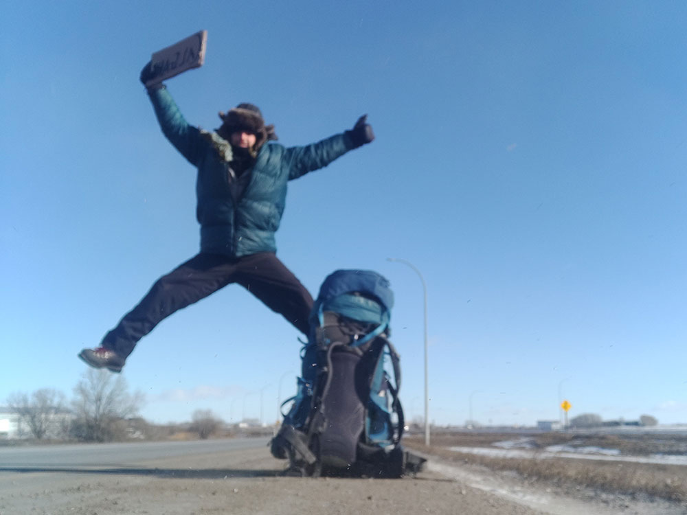 A man holding a cardboard sign by the side of the road jumps into the air, giving a thumbs up. He is wearing winter gear, the side of the road is snowy, and he’s beside a large backpack.